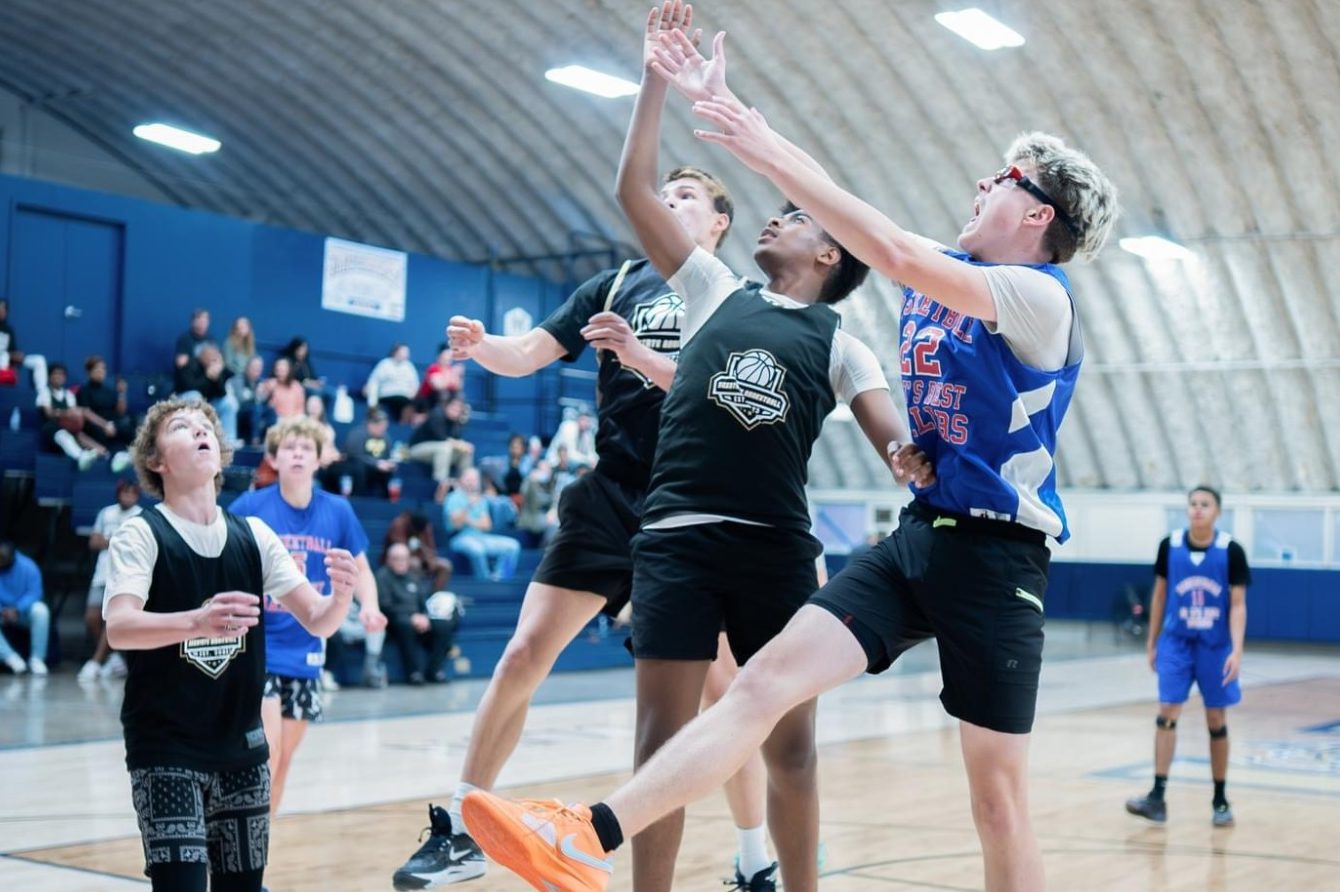 Two boys jump to block a basketball shot during a game, with spectators in the background.